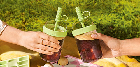 Two hands clinking iced drinks in reusable jars at an outdoor picnic.
