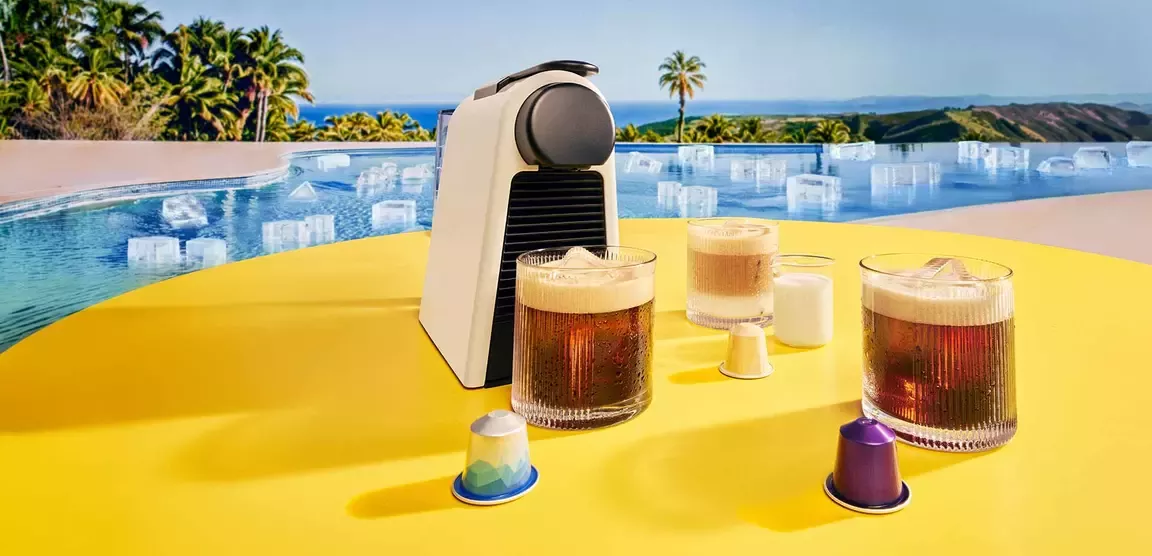 Nespresso coffee drinks and capsules on a yellow table by a pool overlooking a tropical landscape.