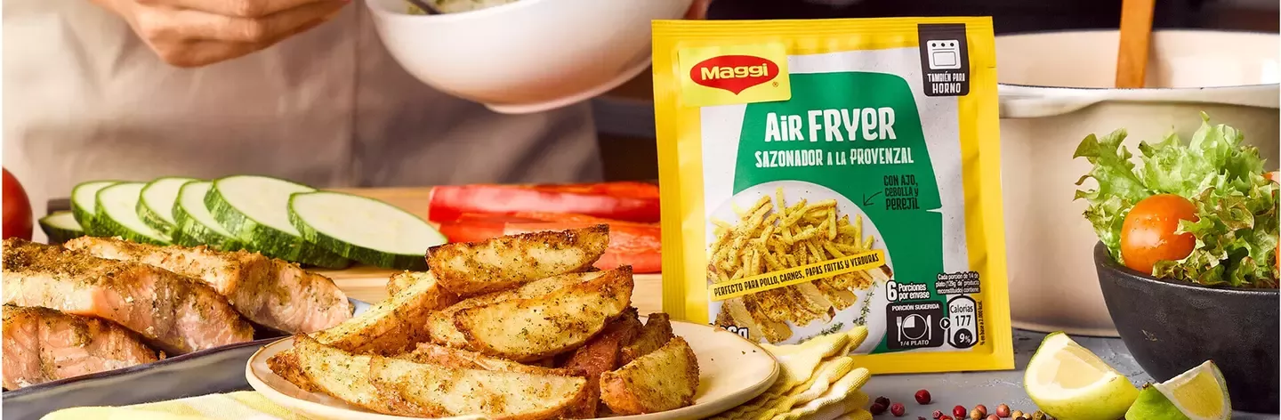 A person preparing food with a bowl of seasoning, alongside air-fried potato wedges, vegetables, and Maggi seasoning packet.