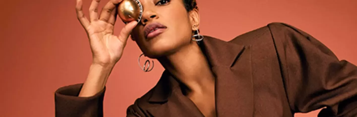 Person in a brown blazer holding a small coffee pod to one eye against a warm-toned background.