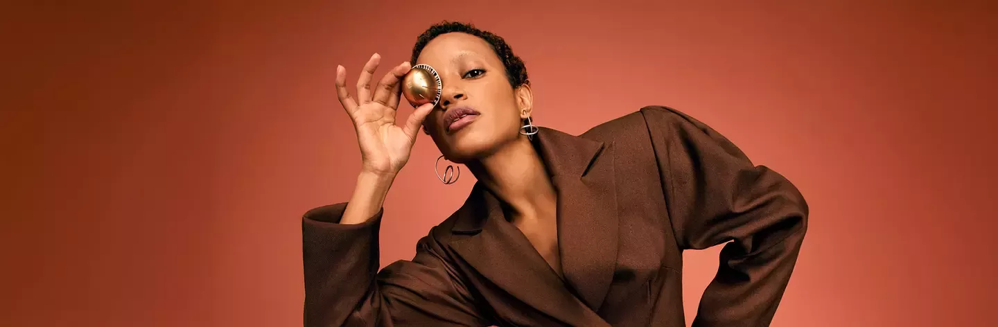 Person in a brown blazer holding a small coffee pod to one eye against a warm-toned background.