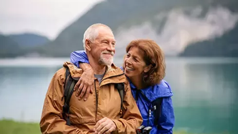 Older couple hiking together by a mountain lake wearing outdoor jackets and backpacks