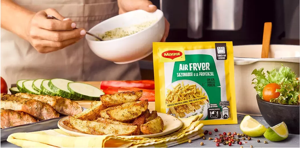 A person preparing food with a bowl of seasoning, alongside air-fried potato wedges, vegetables, and Maggi seasoning packet.