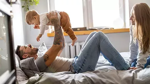 man laying on bed and is holding his son, his wife is sitting next to him