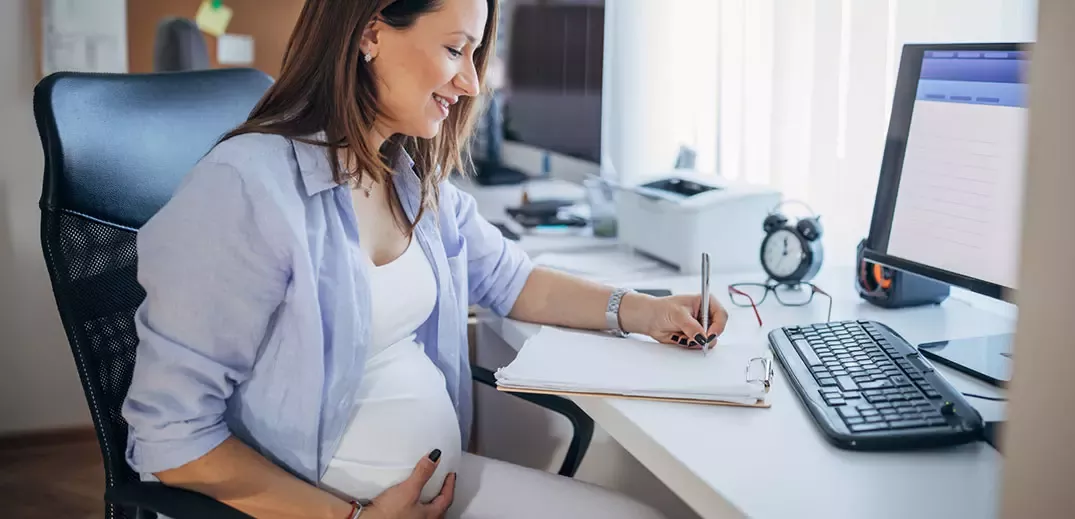 Pregnant employee writing notes at office desk with computer and keyboard