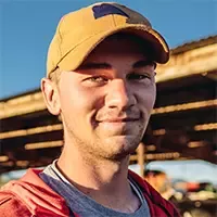 Person wearing a red hoodie and tan cap standing in a cattle farm with wooden shelter