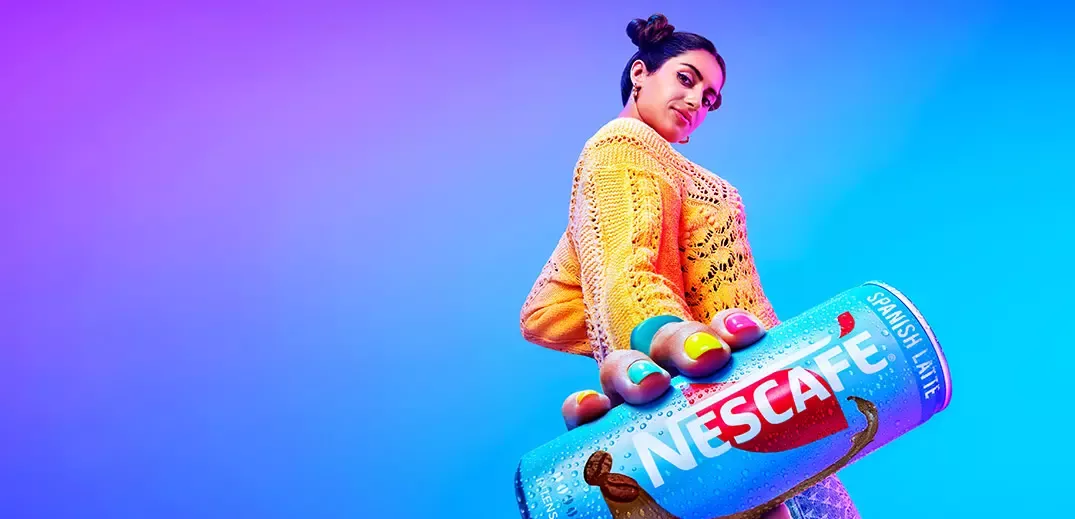 Person holding a blue Nescafé Spanish Latte can against a vibrant blue‑purple background.