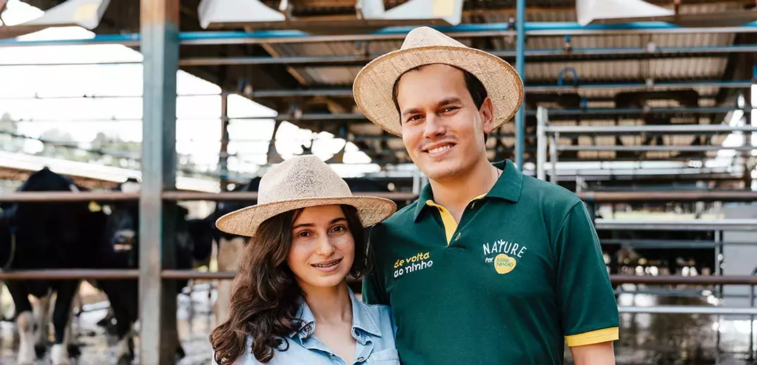 Two people wearing straw hats standing in a dairy farm with cows and feeding structures behind